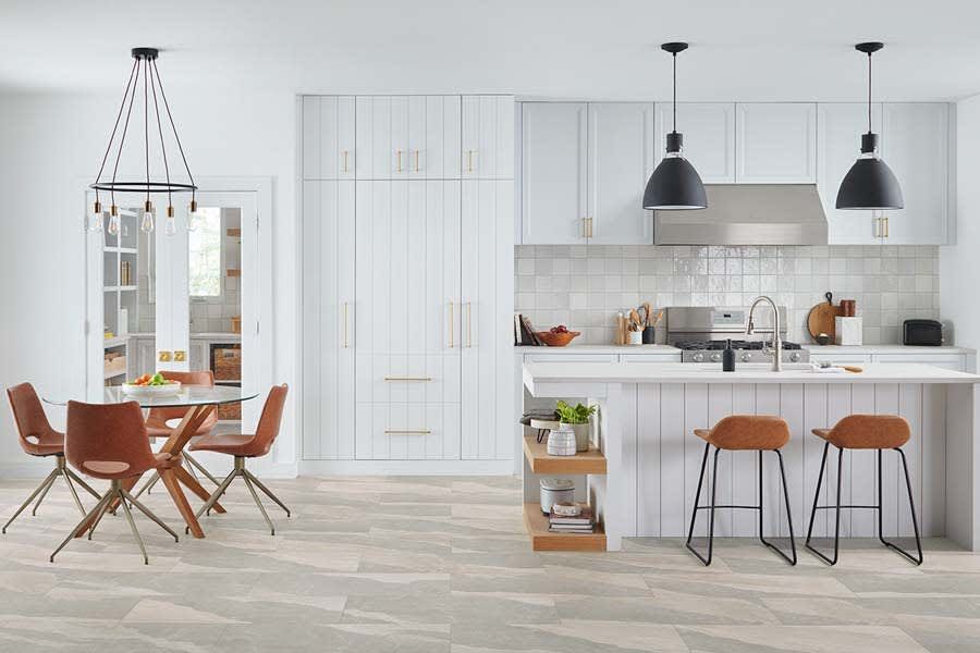 A modern white kitchen with a dining area, a large island with two stools, and a tall pantry unit on light wood floors.