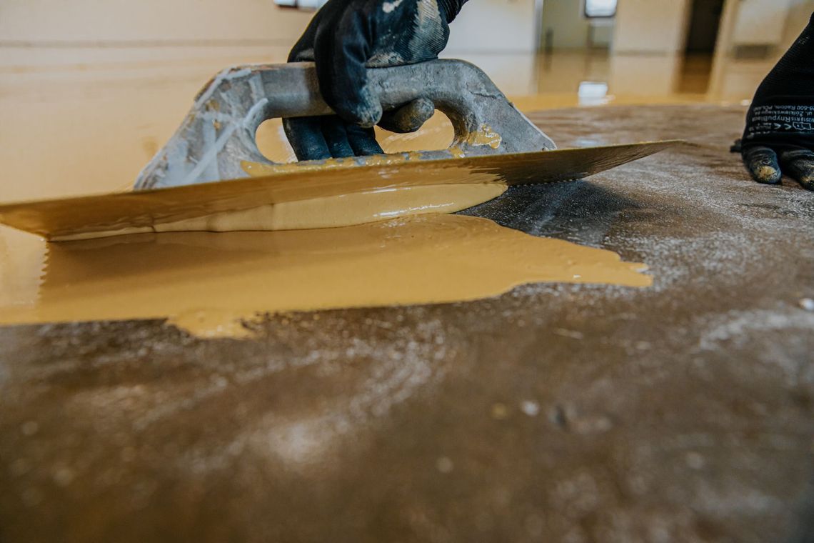 A hand trowel smoothing brown paste on a surface beside a blue-gloved hand