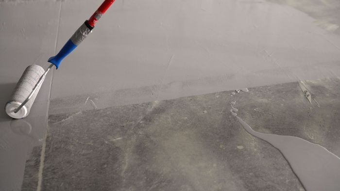 Paint roller applying light gray paint to a wall corner with a red and blue handle.