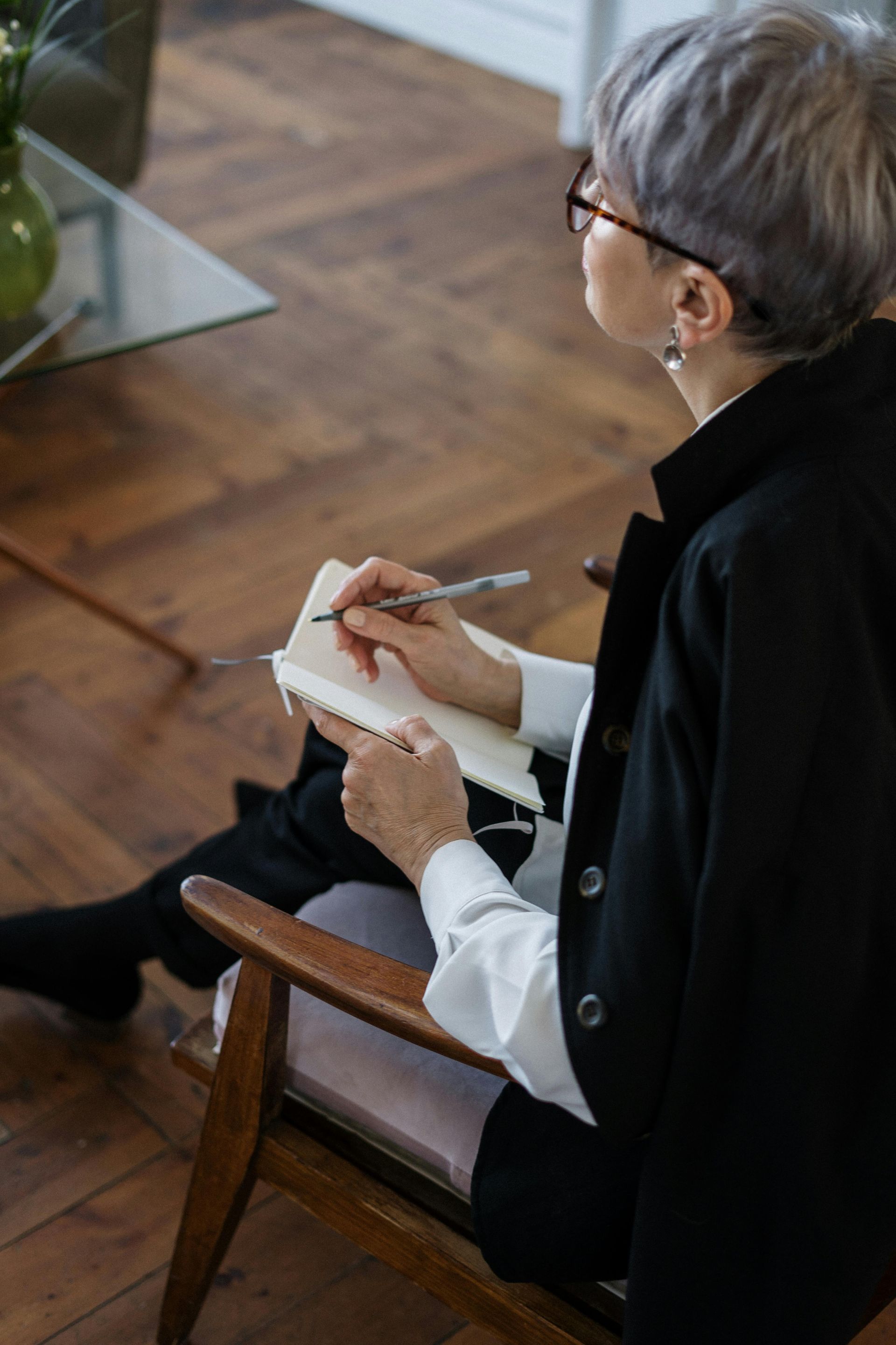 Woman with gray hair writing in a notebook, sitting in a wooden chair, indoors.