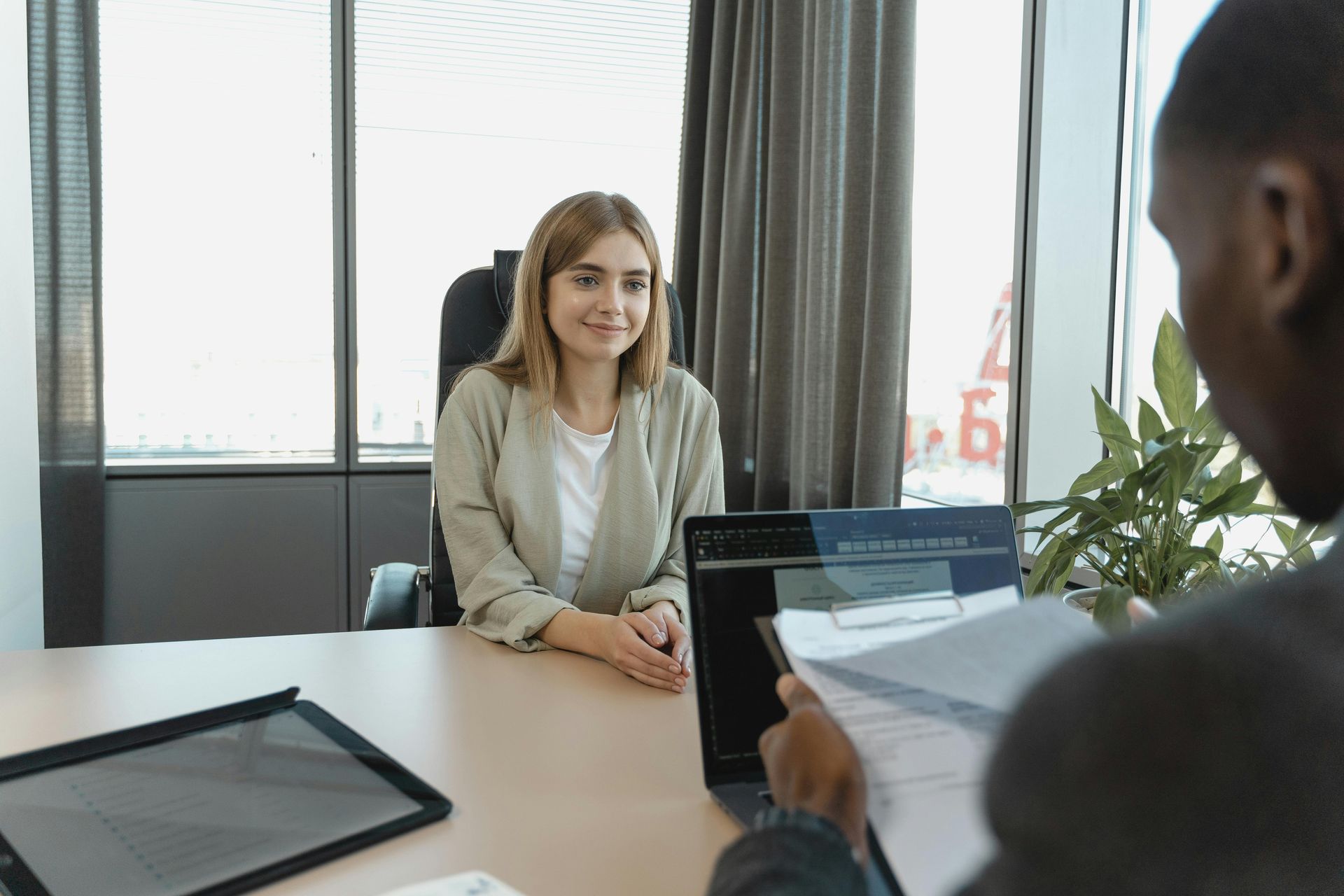 Woman seated at a desk, smiling at a person holding papers and a laptop in a modern office.