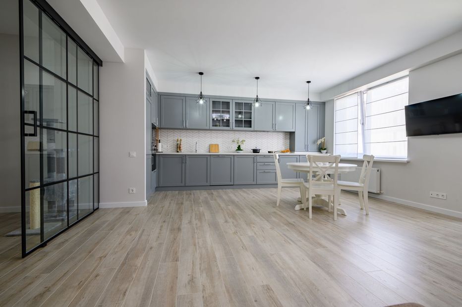 Bright modern kitchen and dining area with gray cabinets, white table, and large windows in an open room