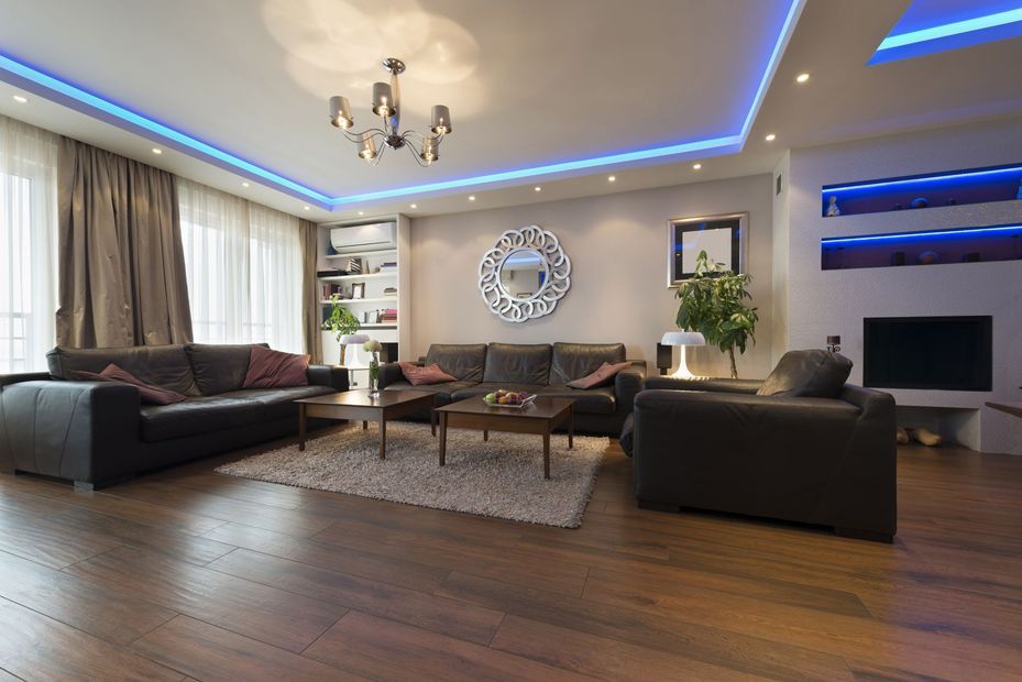 Modern living room with sofas, hardwood floors, blue ceiling lights, and a TV on the wall