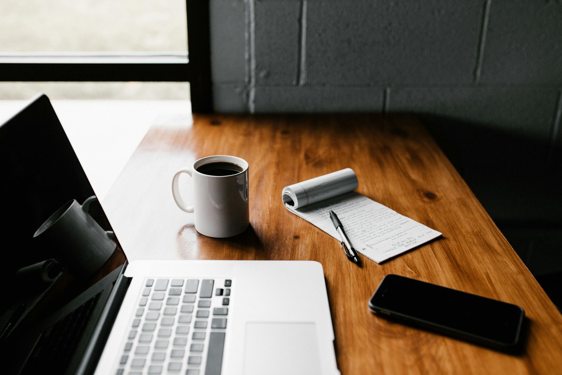 A wooden desk with a laptop , a cup of coffee , a notebook , and a cell phone getting ready to create an itinerary