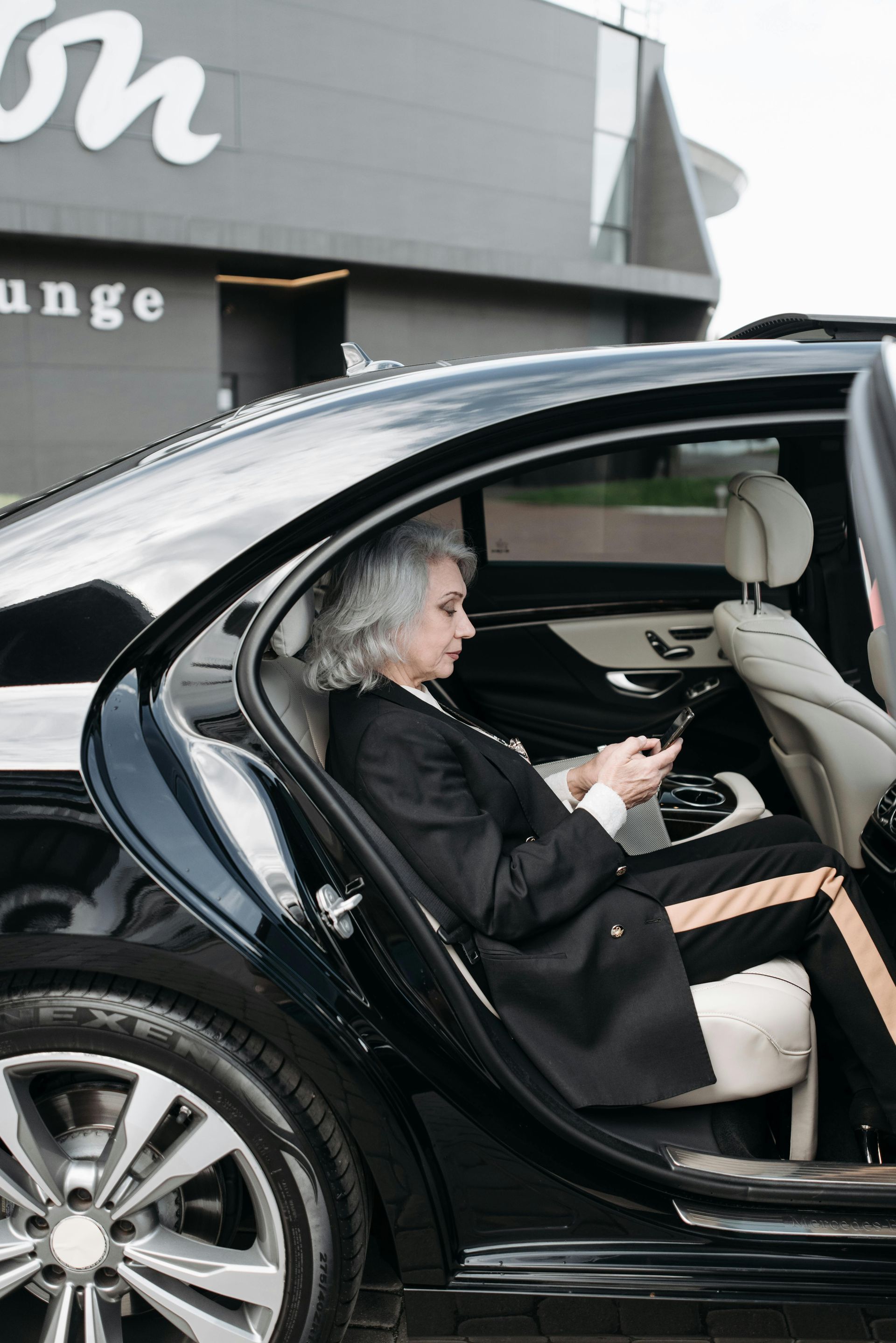 Person in a black car, using a phone. The car door is open. In the background, there's a building with the word