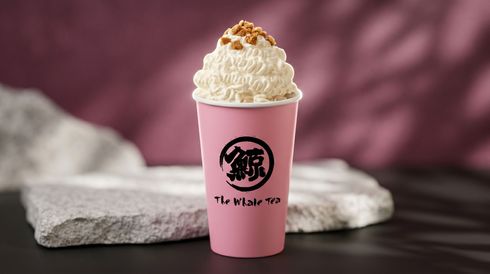 Pink cup with a black logo, topped with whipped cream and caramel, on a dark tabletop with a stone backdrop