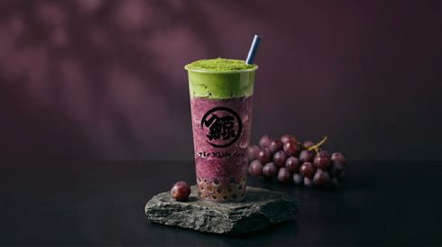 Purple drink in a clear cup with green foam and a straw, beside grapes on a dark surface