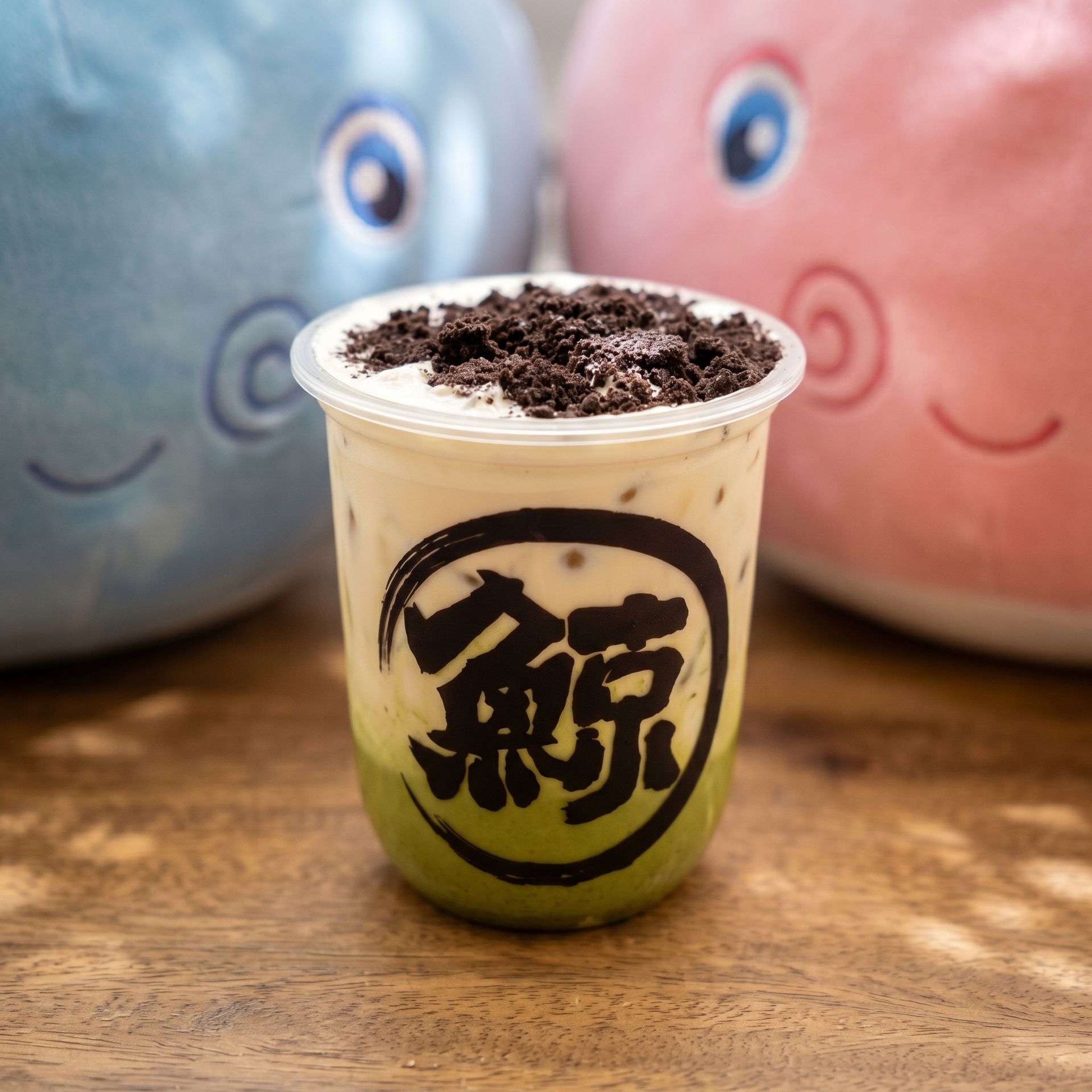 Cup of layered green and black drink topped with crushed crumbs, with two smiling plush toys in the background