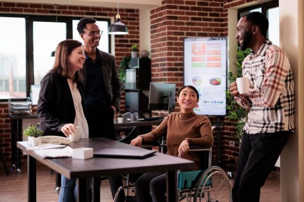 A group of people are standing around a table with a woman in a wheelchair.