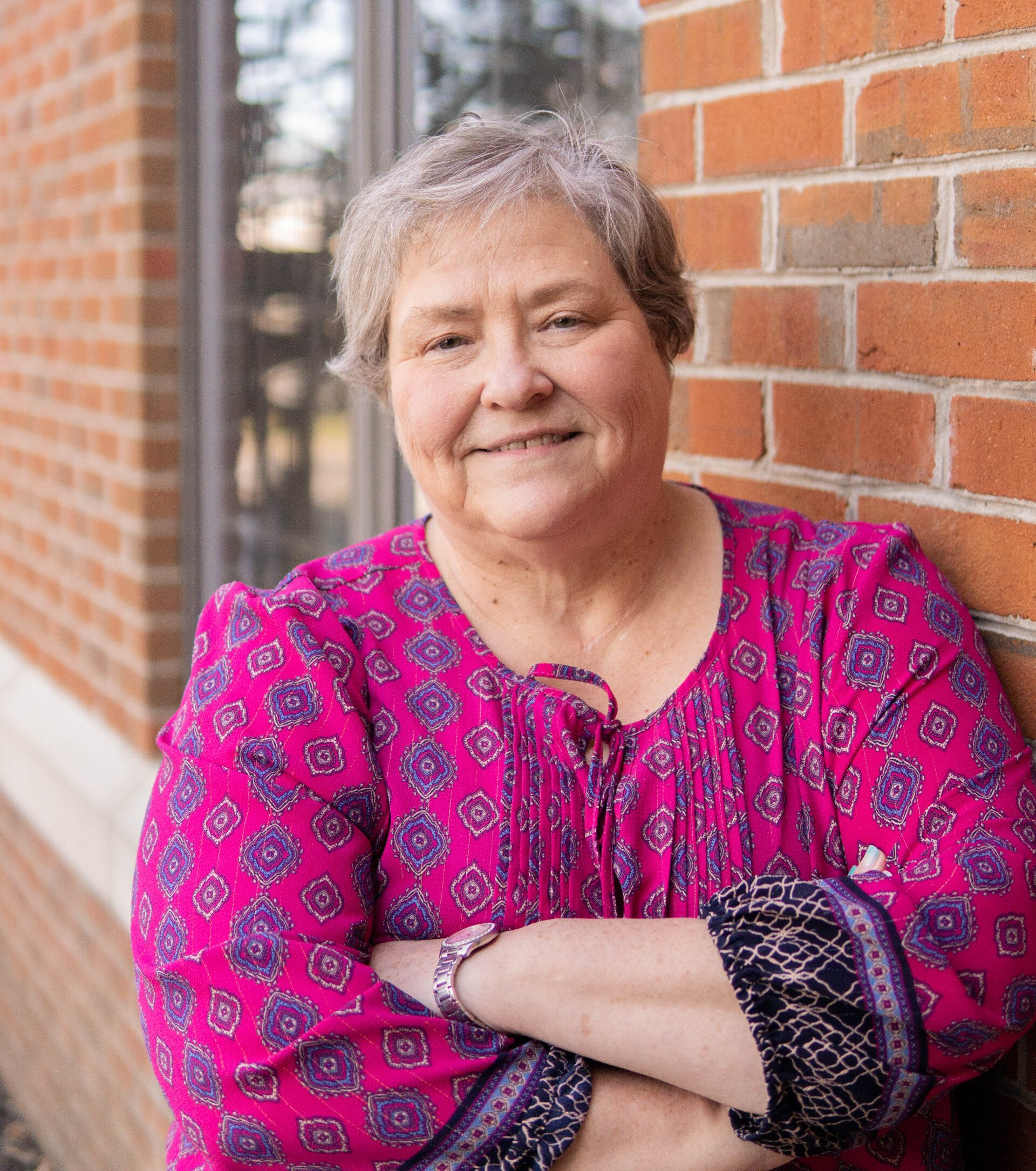 Woman with short gray hair, pink blouse, smiling against a brick wall.