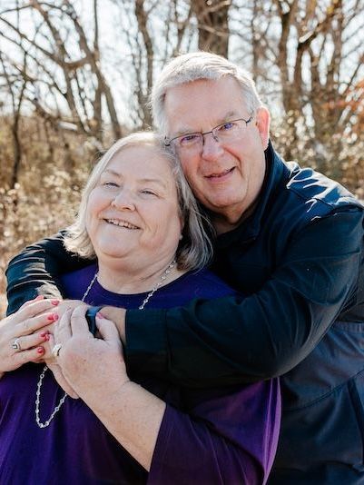Smiling older couple embracing outdoors; man's arms around woman, both wearing purple, trees in background.