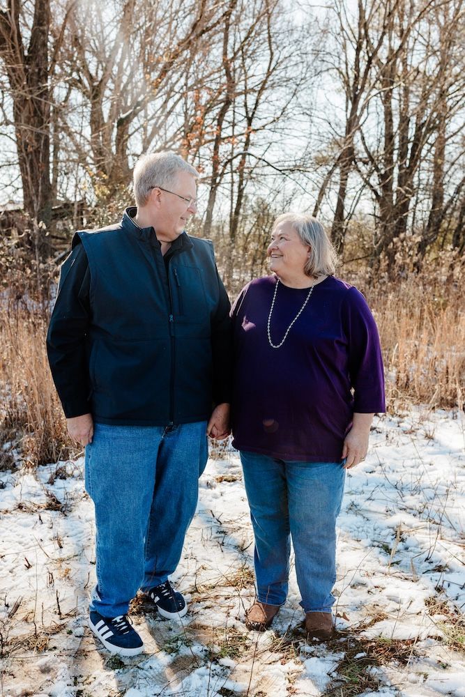 Older couple holding hands, smiling at each other. Man in black vest, jeans, woman in purple shirt, jeans, snowy field.