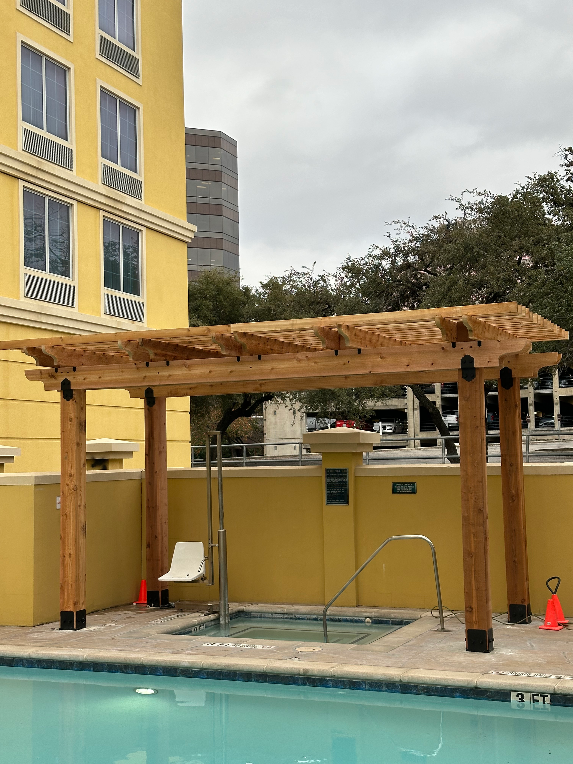 A wooden pergola over a swimming pool with a yellow building in the background