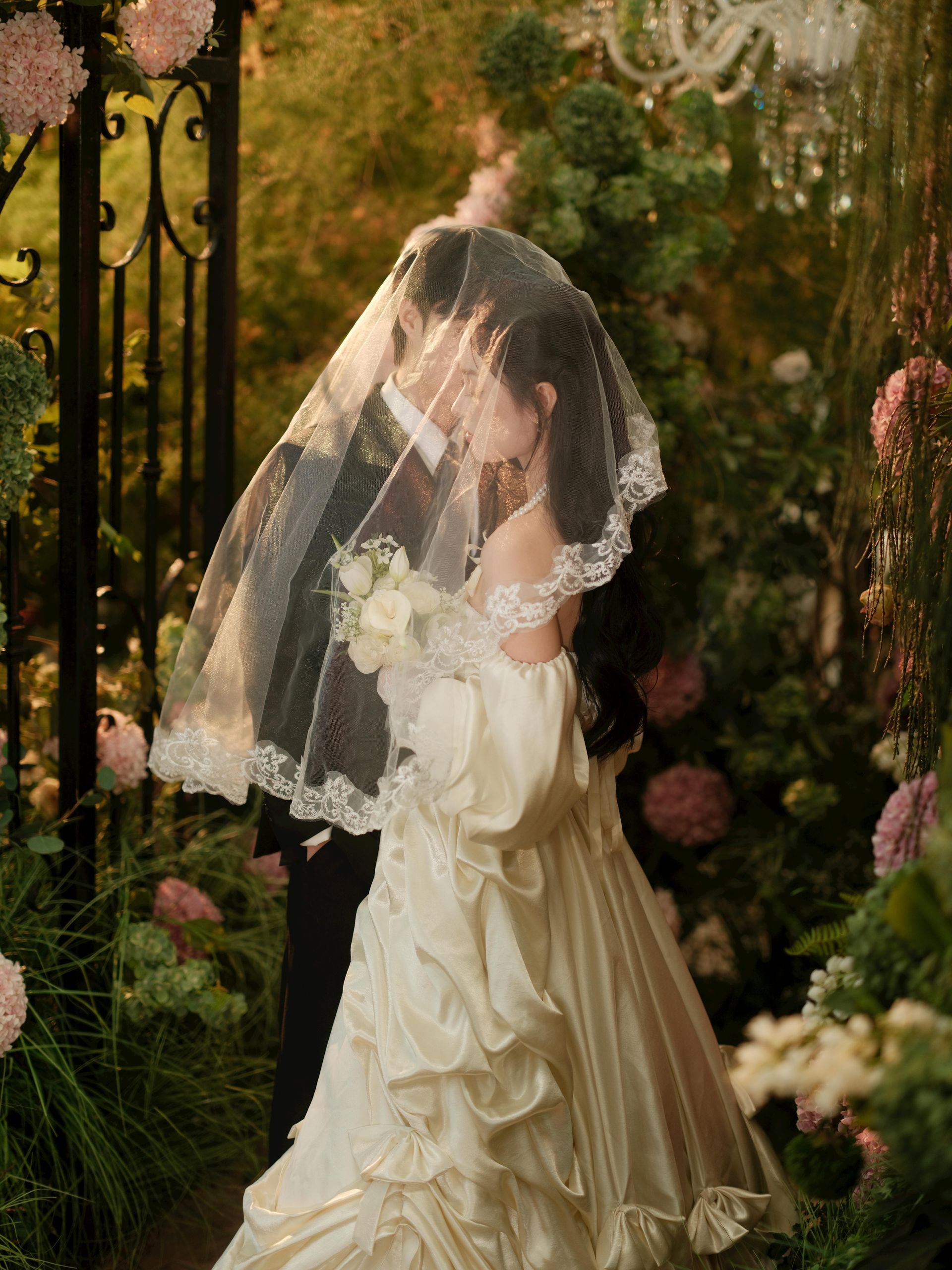 Bride in a cream gown and veil standing in a lush garden by an iron gate.