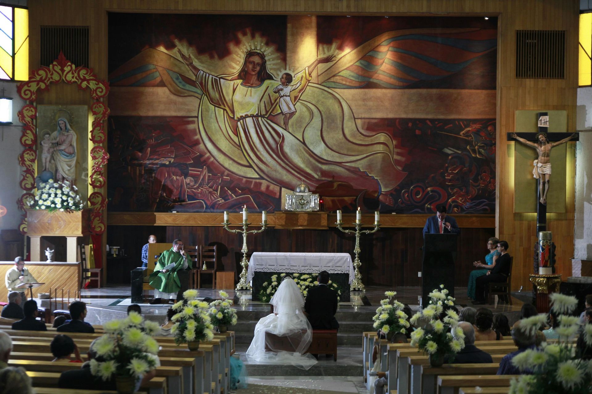 Church altar with a crucifix, large Jesus mural, floral arrangements, and a white-robed figure in the center aisle