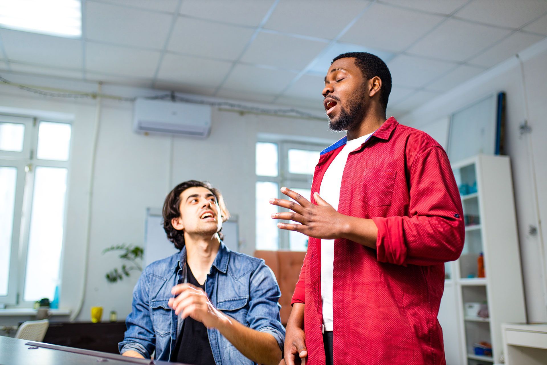 Two coworkers talking in a bright office, one seated and smiling, the other standing in a red shirt and gesturing.