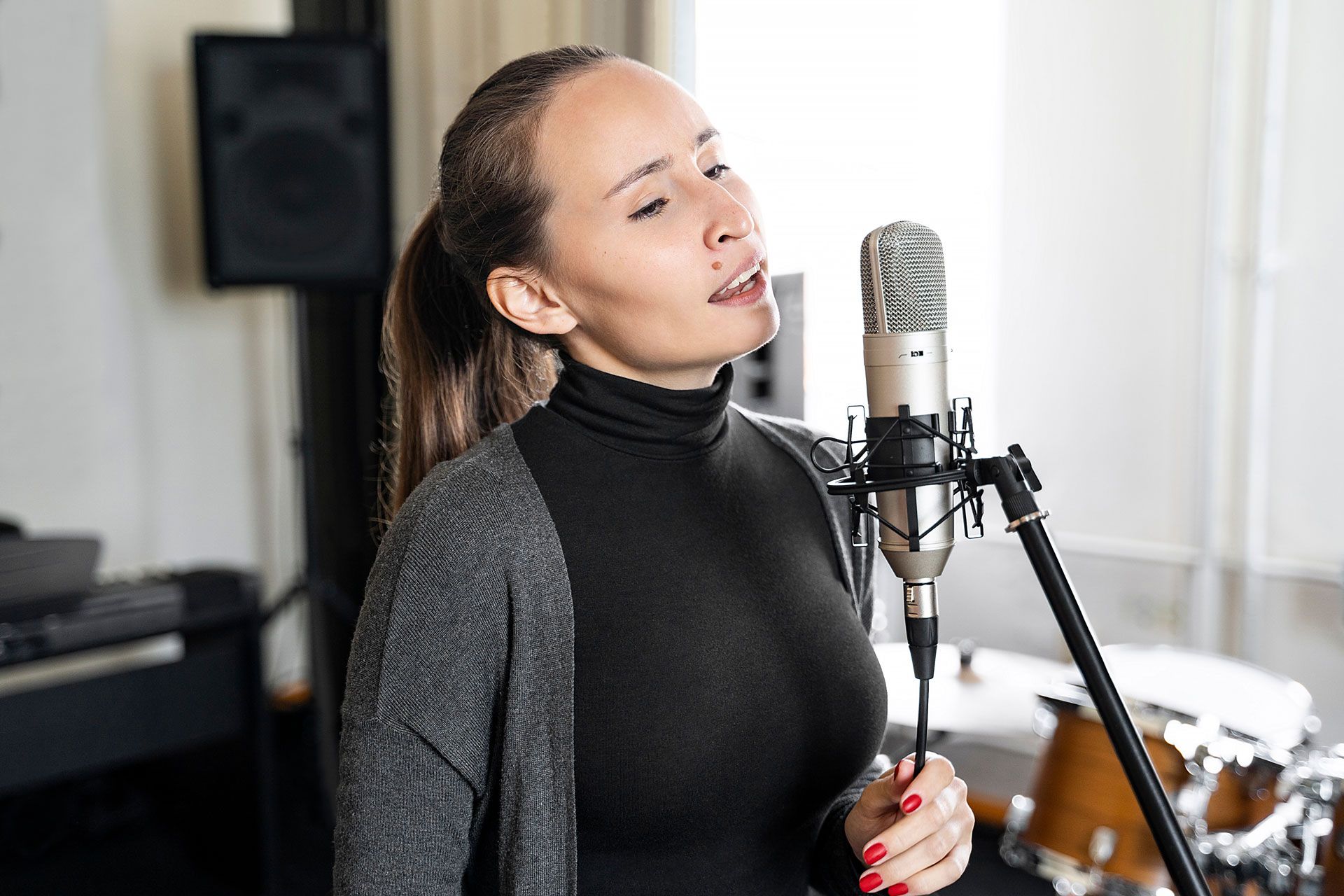 Singer performing into a studio microphone, wearing a black turtleneck and gray cardigan.
