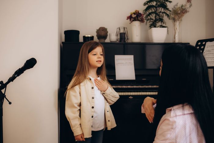 Child standing by piano as a person plays, in a cozy room with plants and a microphone stand.