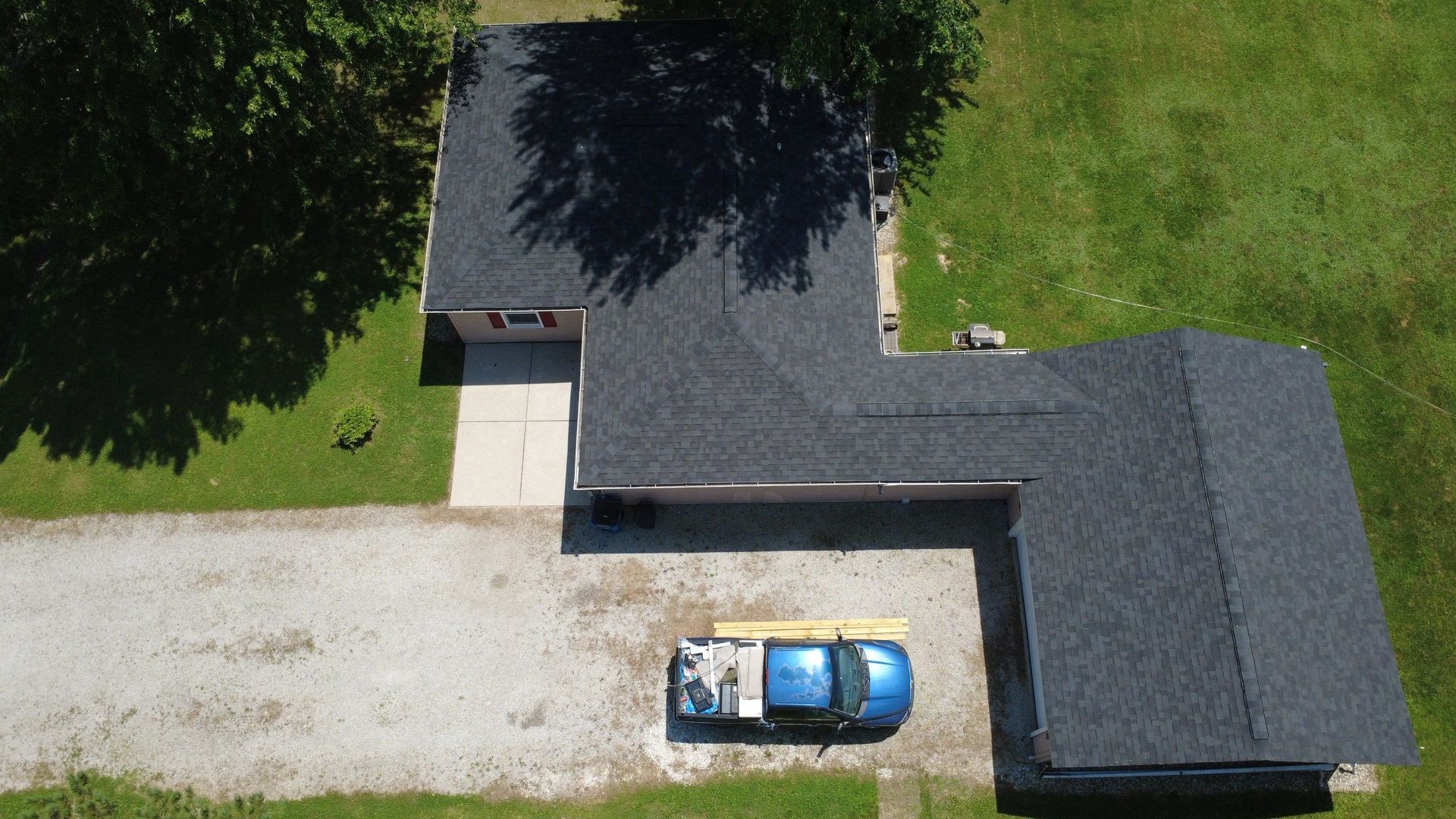 Aerial view of a house with a gravel driveway, a blue car, and a small outbuilding surrounded by green lawn.