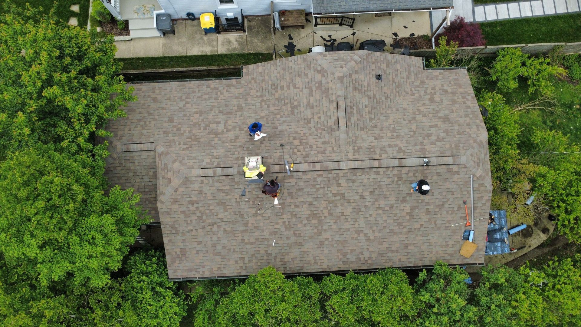 Aerial view of a shingled rooftop with two workers and tools near the center, surrounded by trees and a driveway.