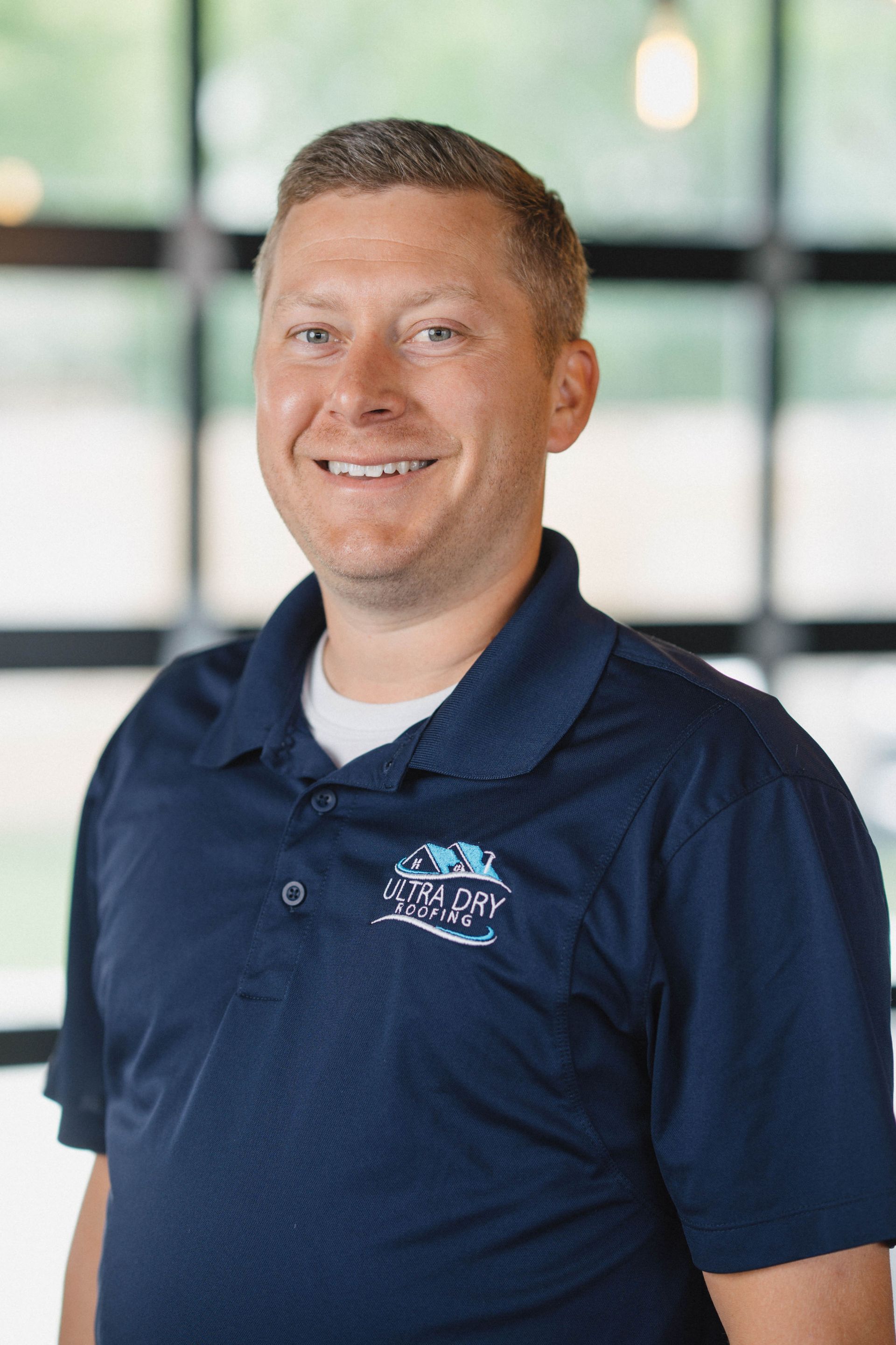 Man in a navy polo shirt smiling in front of a blurred windowed background