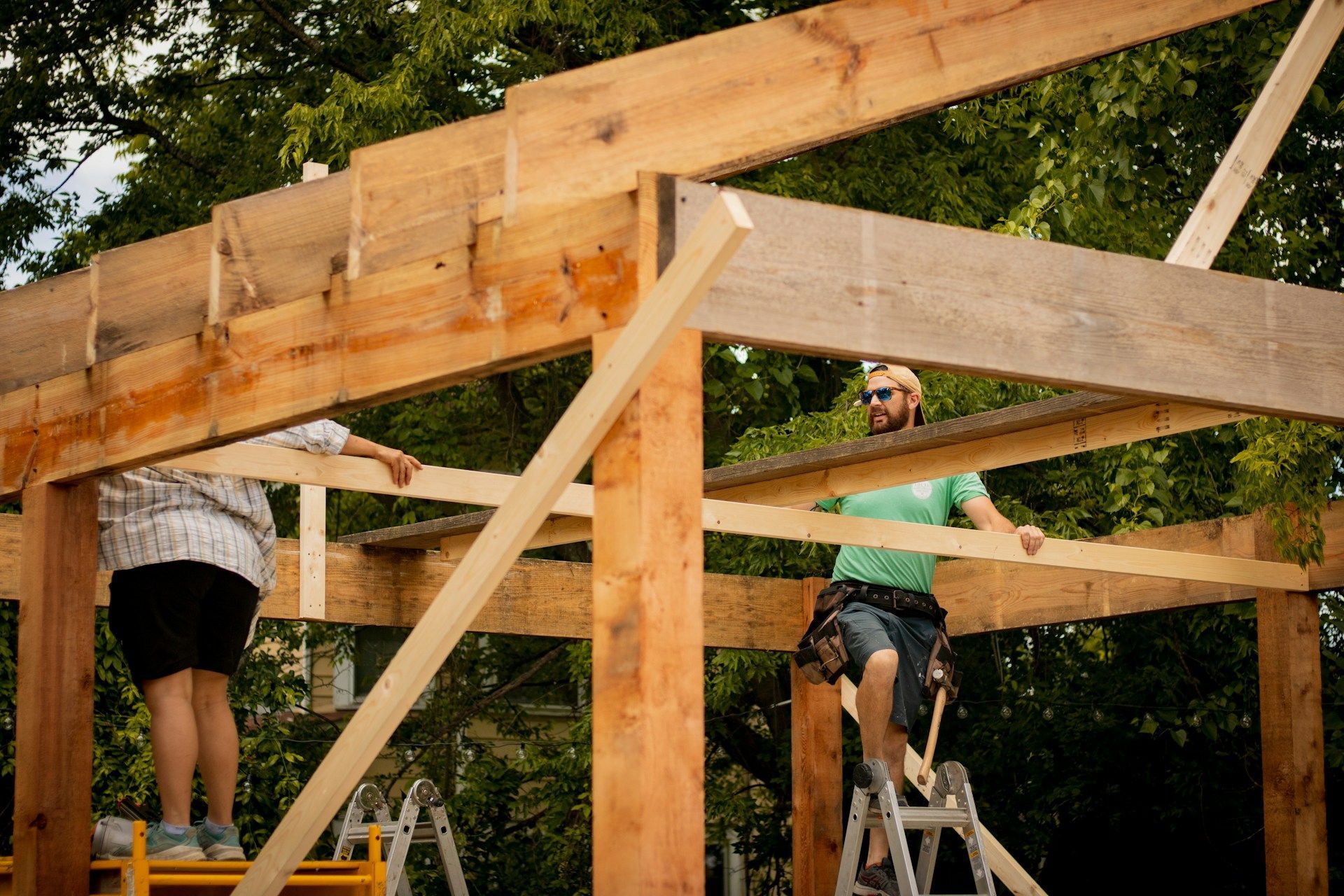 People framing a wooden structure outdoors on ladders under trees.