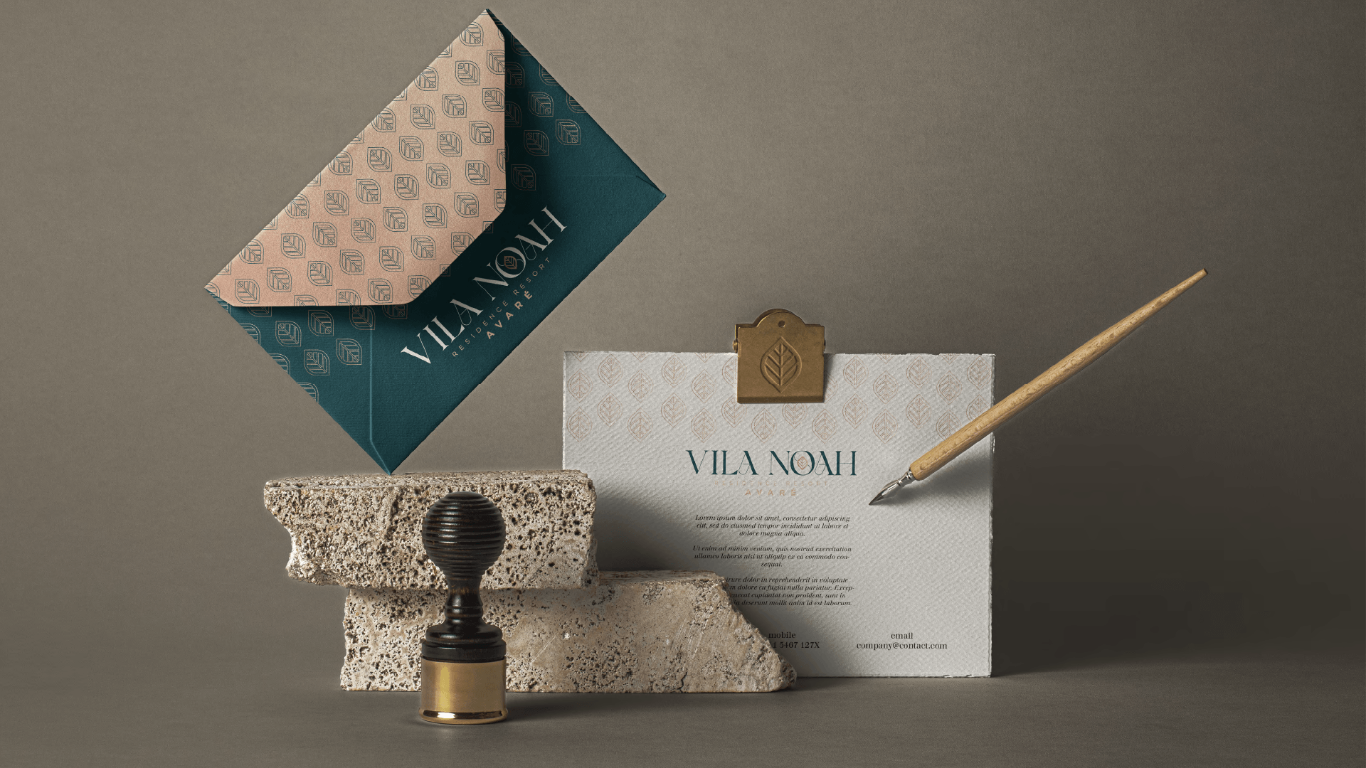 Conjunto de papelaria: um envelope azul-petróleo, um cartão texturizado com um clipe dourado, um selo e uma caneta dispostos sobre uma pedra bruta contra um fundo neutro.
