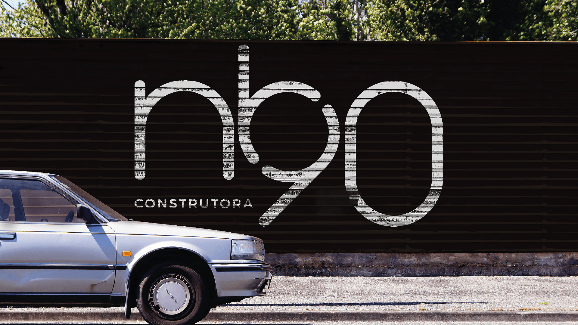 Carro prateado estacionado em frente a um muro preto com grandes placas brancas com as inscrições “nbo” e “constructora”.