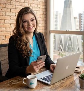 Mulher sorrindo para um laptop em um escritório iluminado, segurando um telefone perto de uma janela com vista para a cidade.