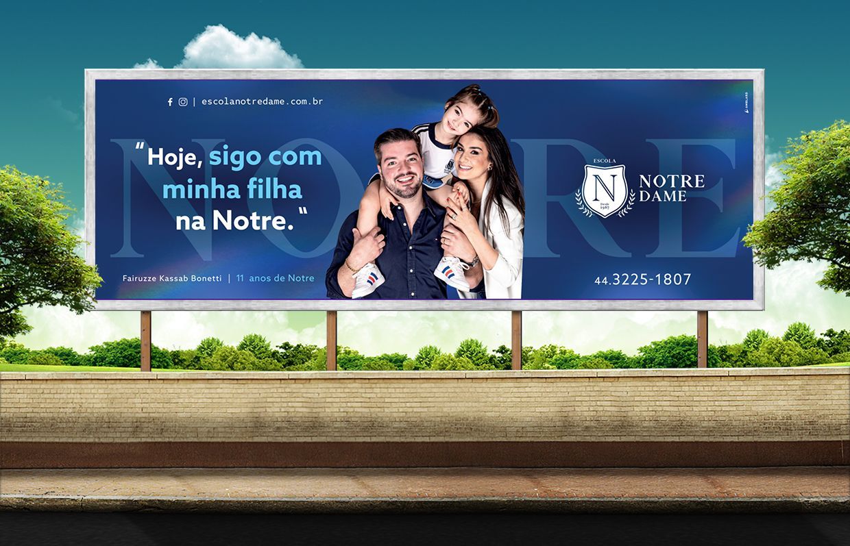 Anúncio em outdoor: Uma família de três pessoas sorri. Um homem e uma mulher abraçam a filha, com o logotipo e o slogan da Notre Dame.
