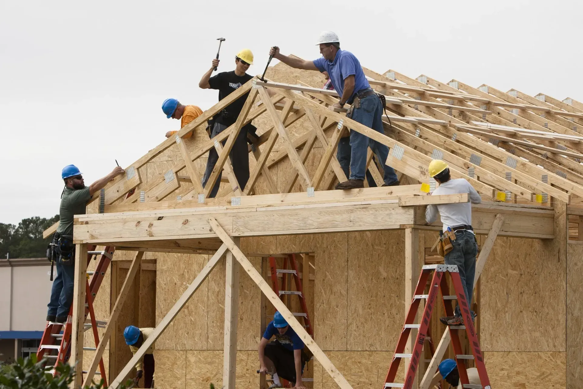 Construction workers on a roof framing a house, using hammers and ladders, under a cloudy sky.