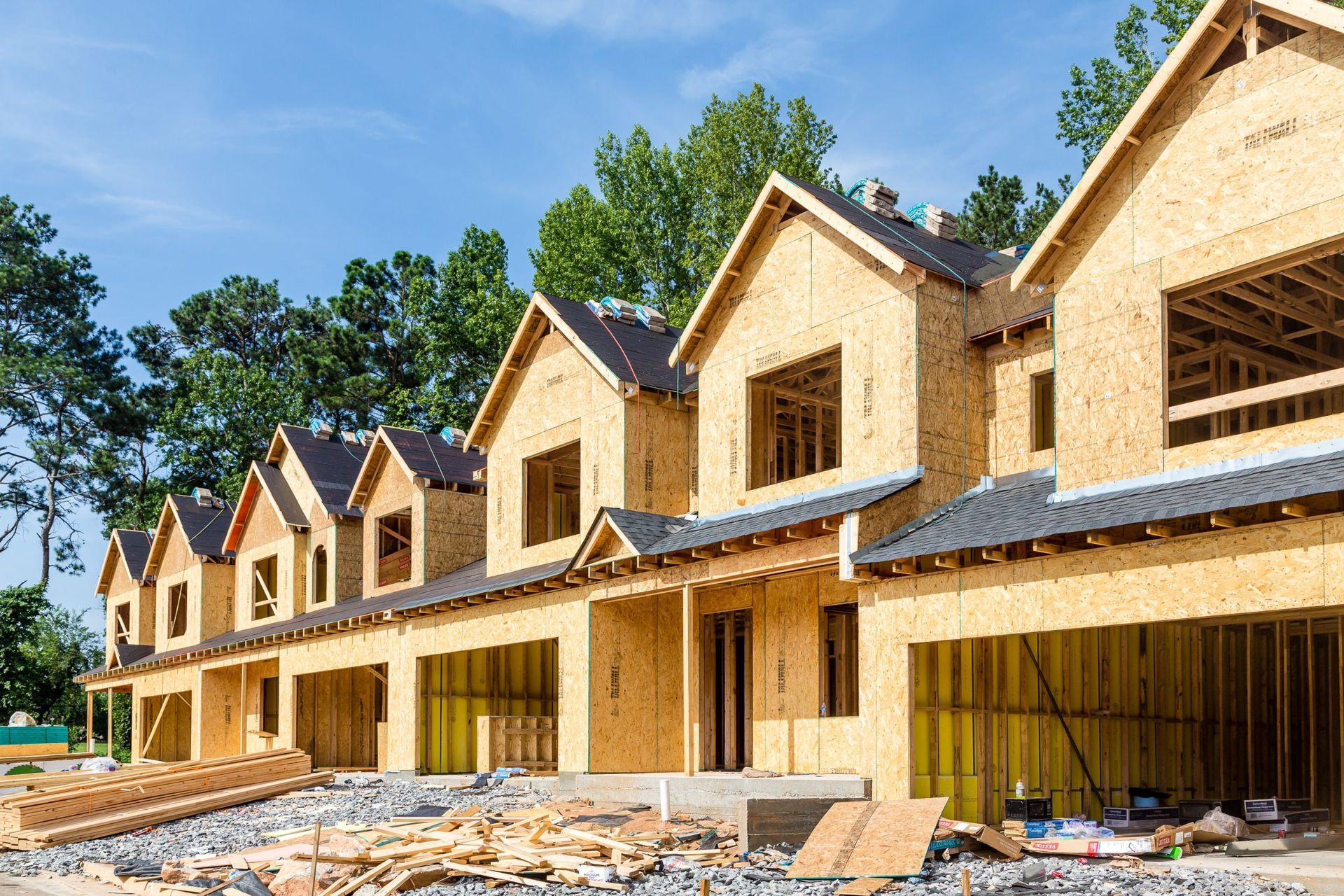 A row of wooden townhouses under construction against a blue sky with green trees in the background.
