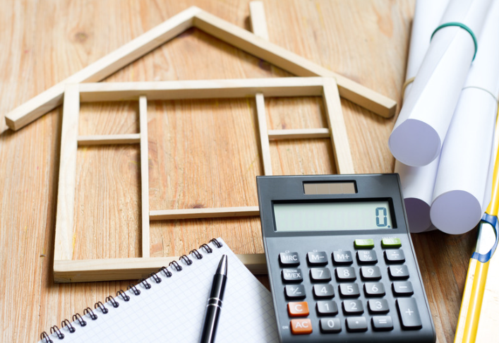 A wooden house model, calculator, notepad with pen, and blueprint rolls on a wooden desk.