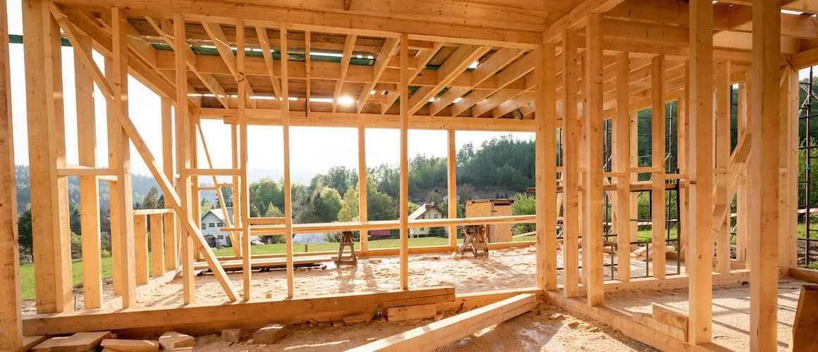 Wooden frame of an unfinished house under construction, looking out at a grassy landscape with distant buildings.