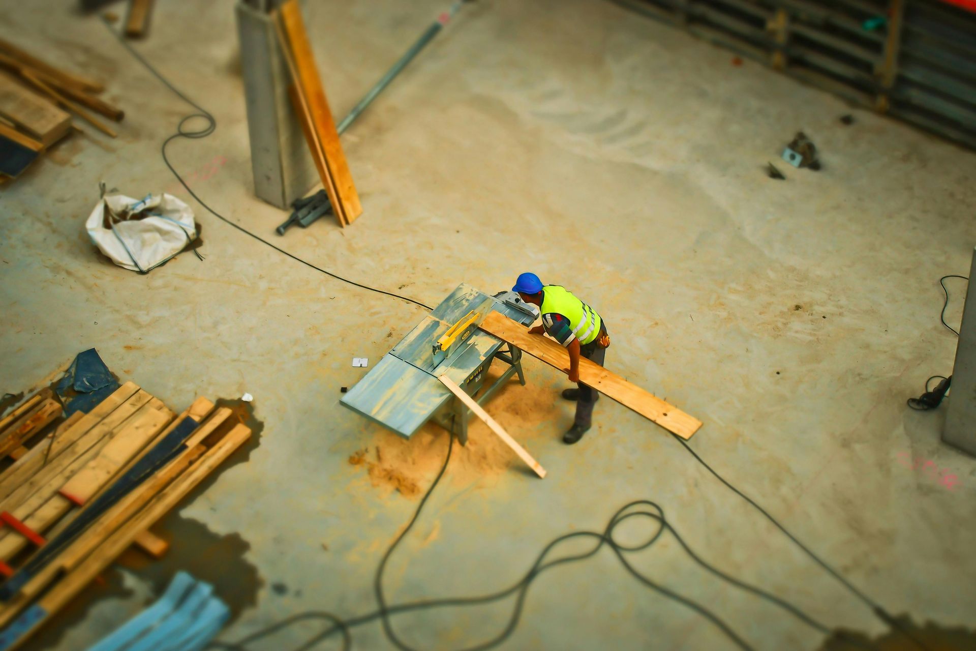 A worker in a high-visibility vest uses a table saw to cut a wooden board on a construction site.