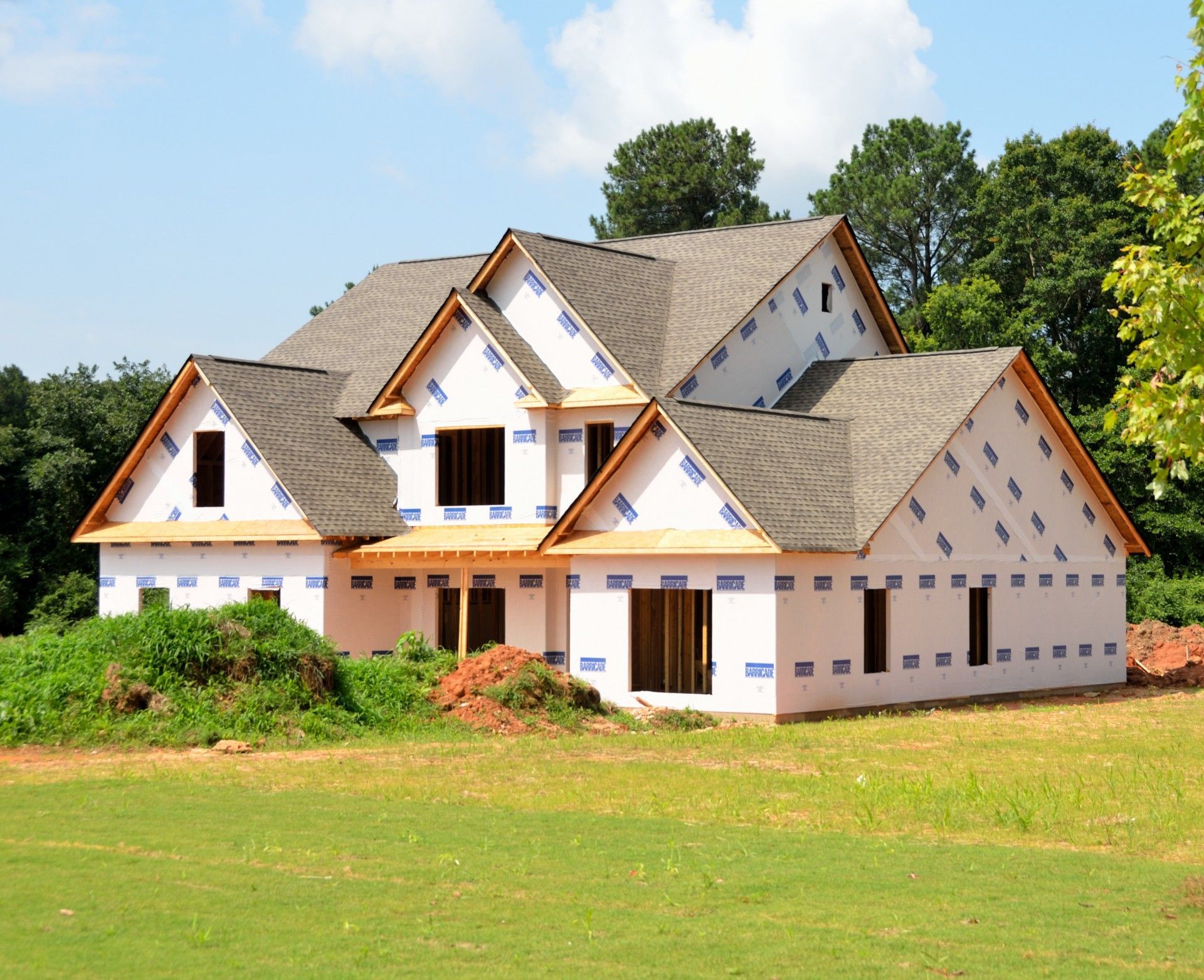 House under construction; roof shingles, wrap, and window frames visible, set in a grassy yard.