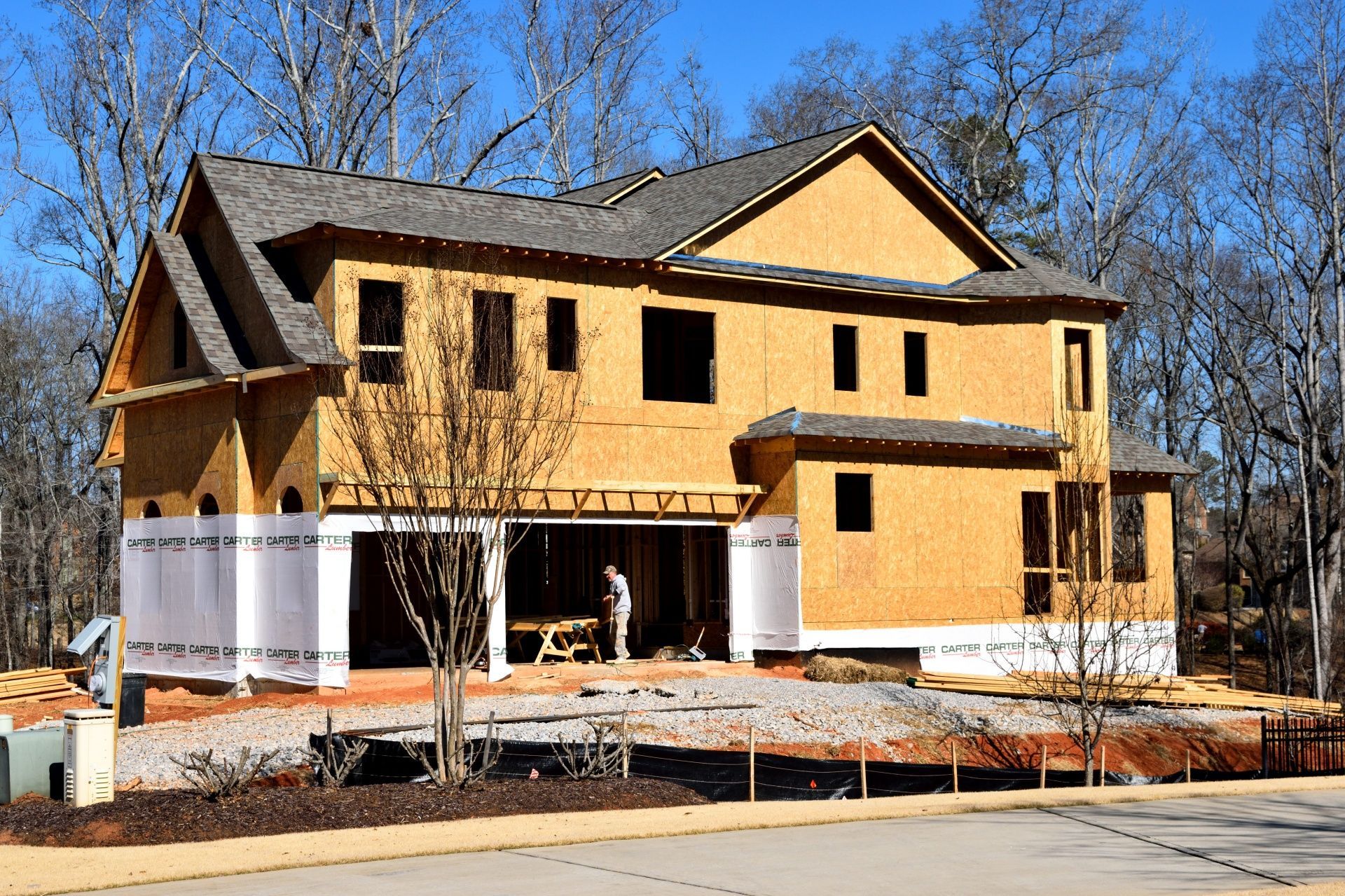 Two-story house under construction, with exposed wooden frame. A worker stands inside.