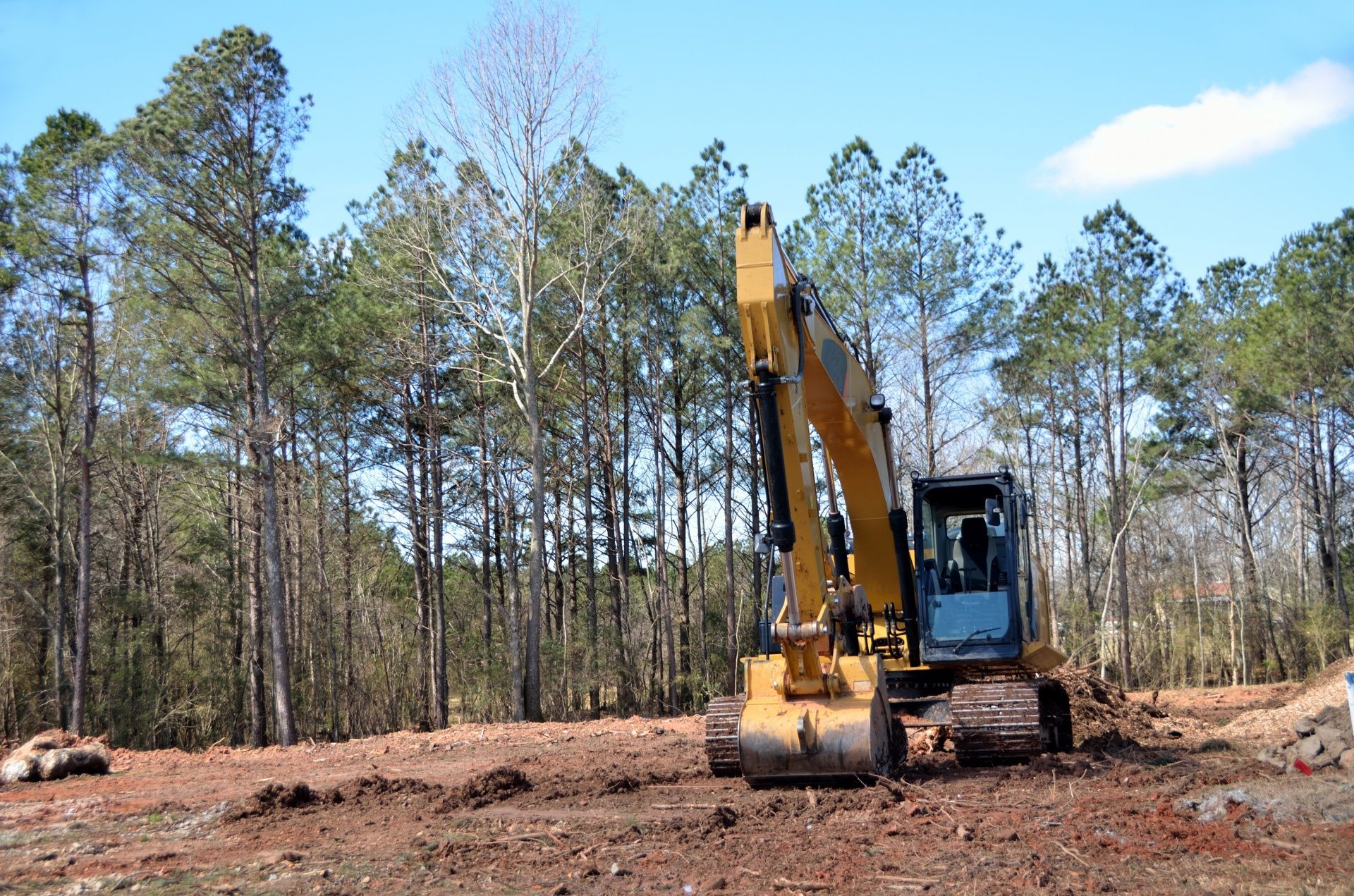 Yellow excavator on a dirt lot, clearing land next to a forest.
