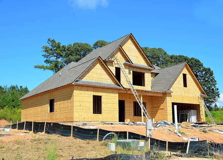House under construction; wood frame, gray roof, ladder, on a hillside under a blue sky.