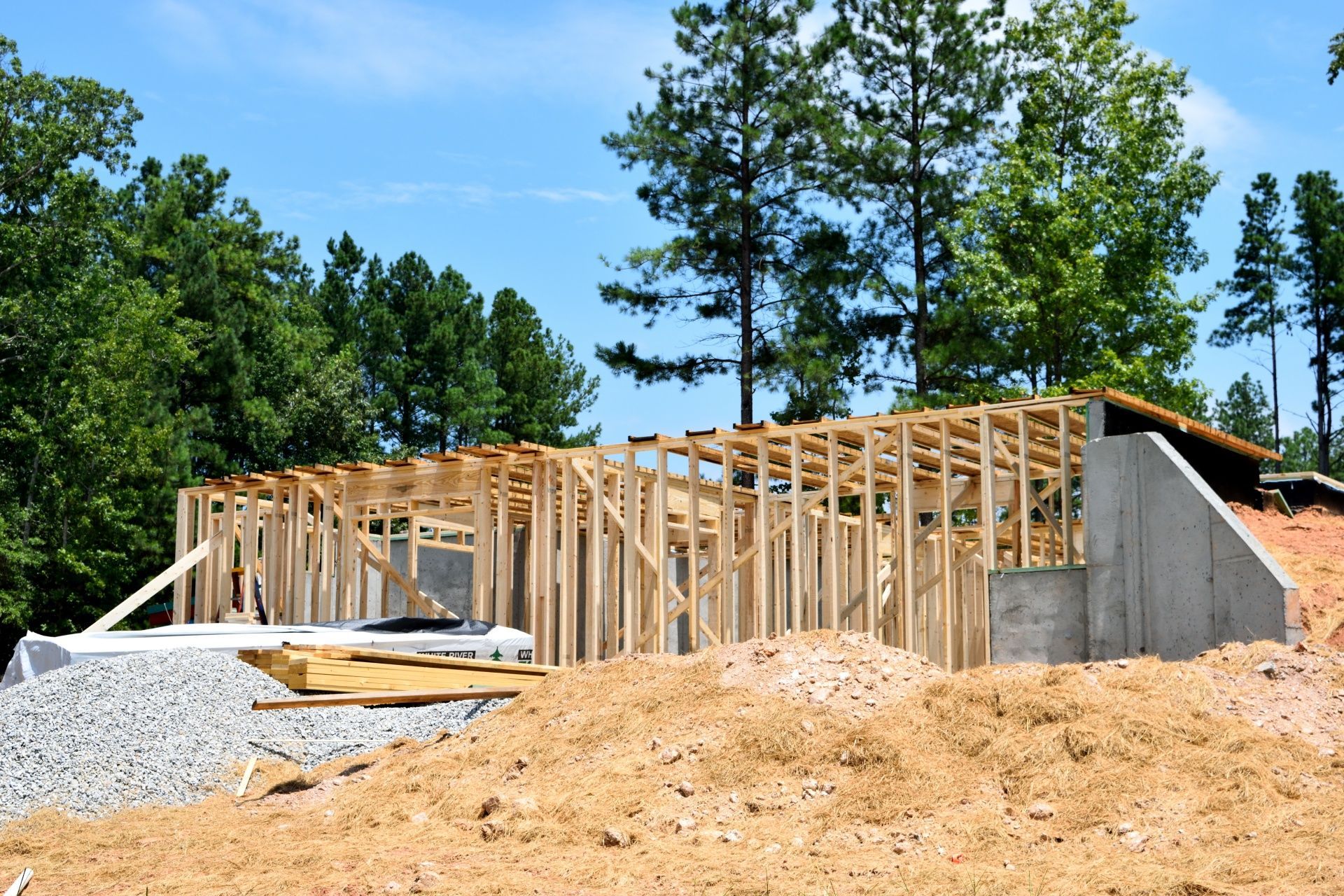 Wooden frame of a building under construction on a dirt and gravel lot, with trees in the background.