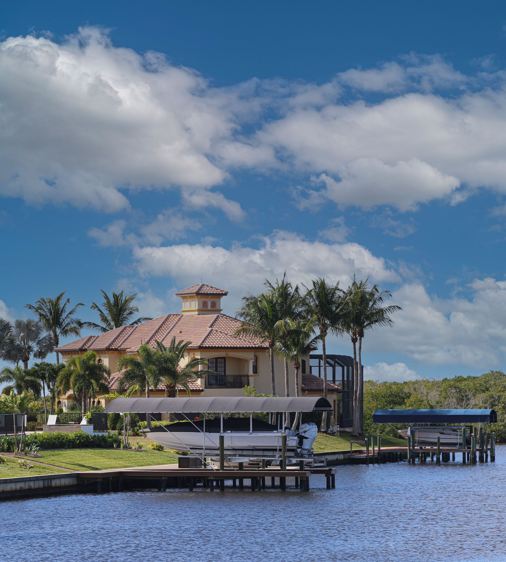 Waterfront house with a red roof, palm trees, and docks beside calm blue water under a cloudy sky