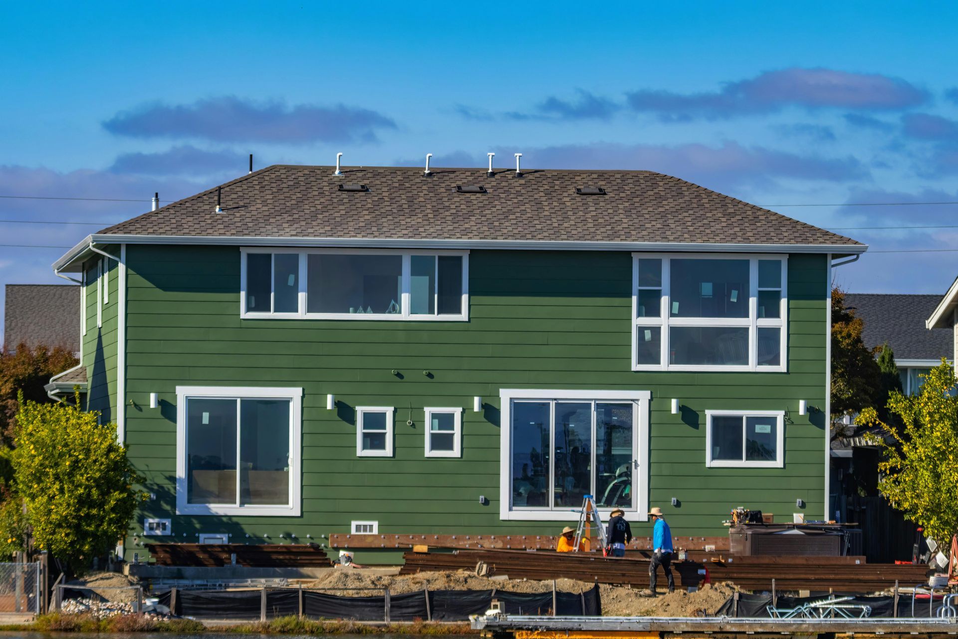 Green two-story house with white trim by the water, with a few people on the deck and rooftop solar panels