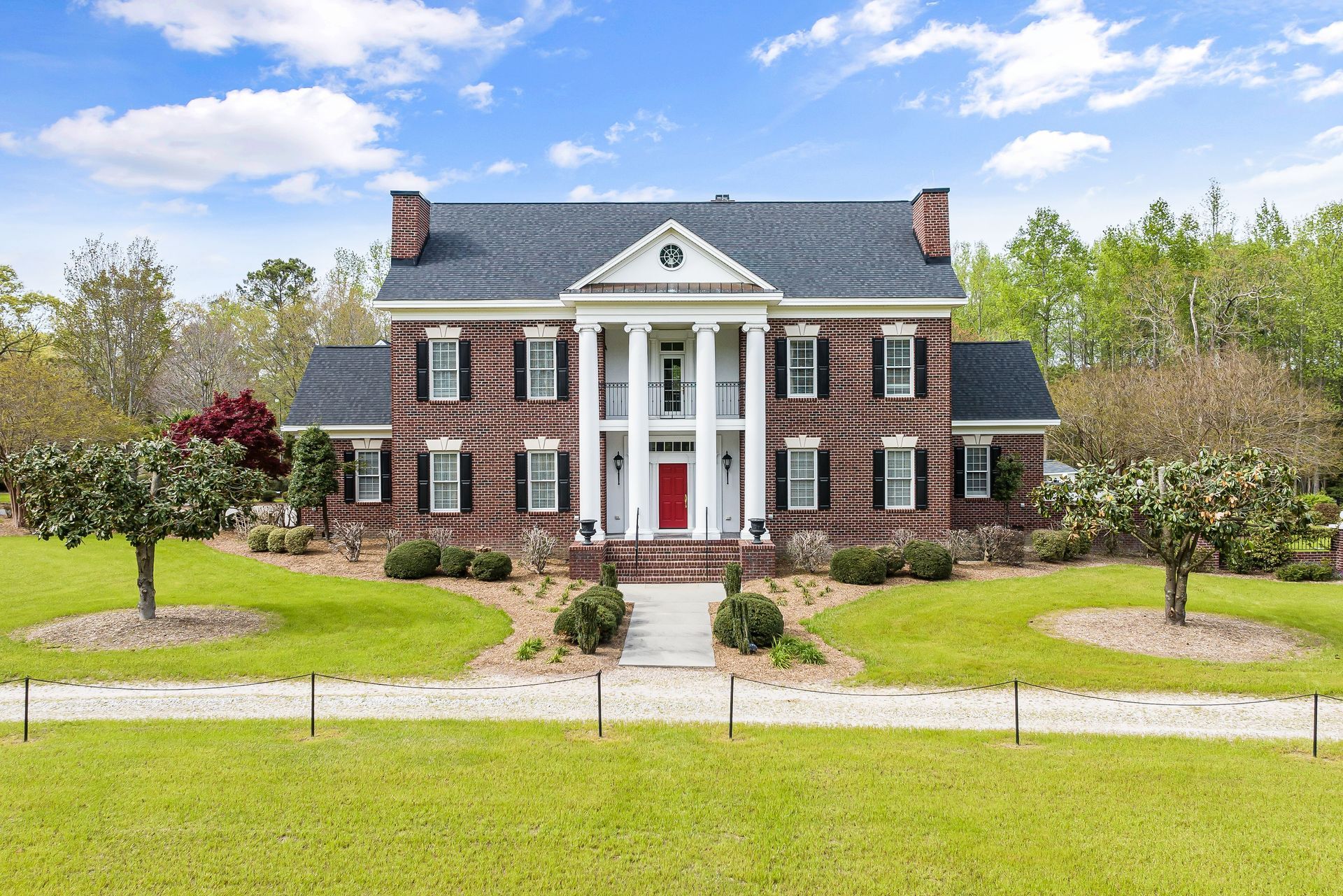 Large colonial-style brick mansion with white columns, red front door, and a manicured lawn under a blue sky