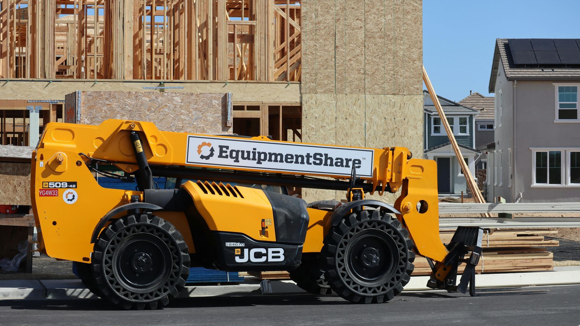 A yellow JCB telescopic forklift parked at a construction site with houses in the background.