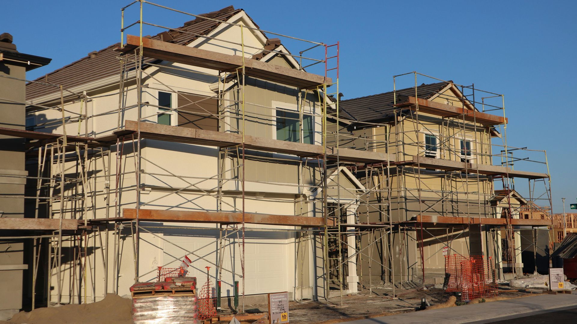Two-story houses under construction with scaffolding. Beige siding, brown roof, and white garage door. Clear blue sky.
