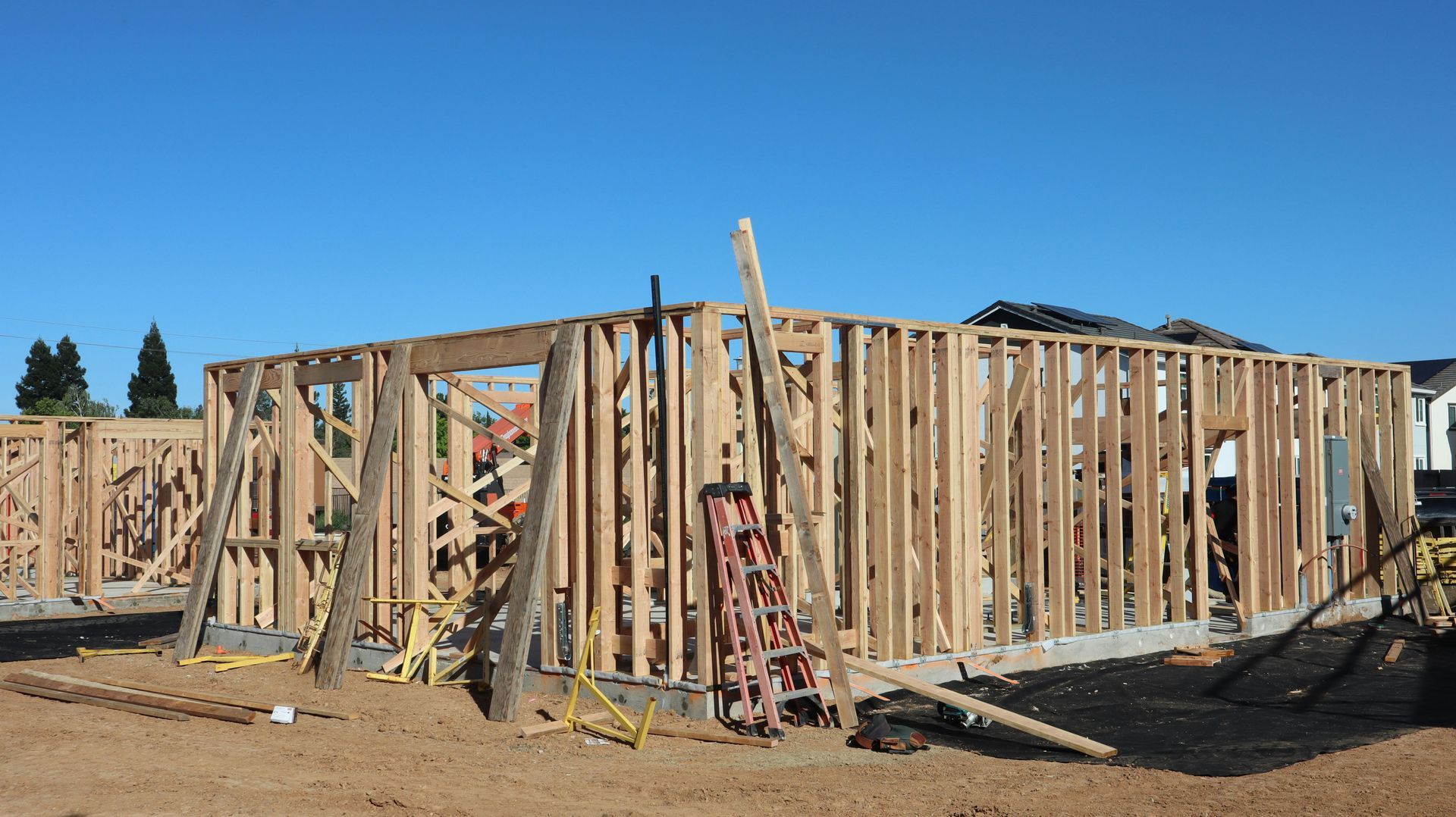 Wood-framed house under construction on a dirt lot, with ladders and scaffolding visible.
