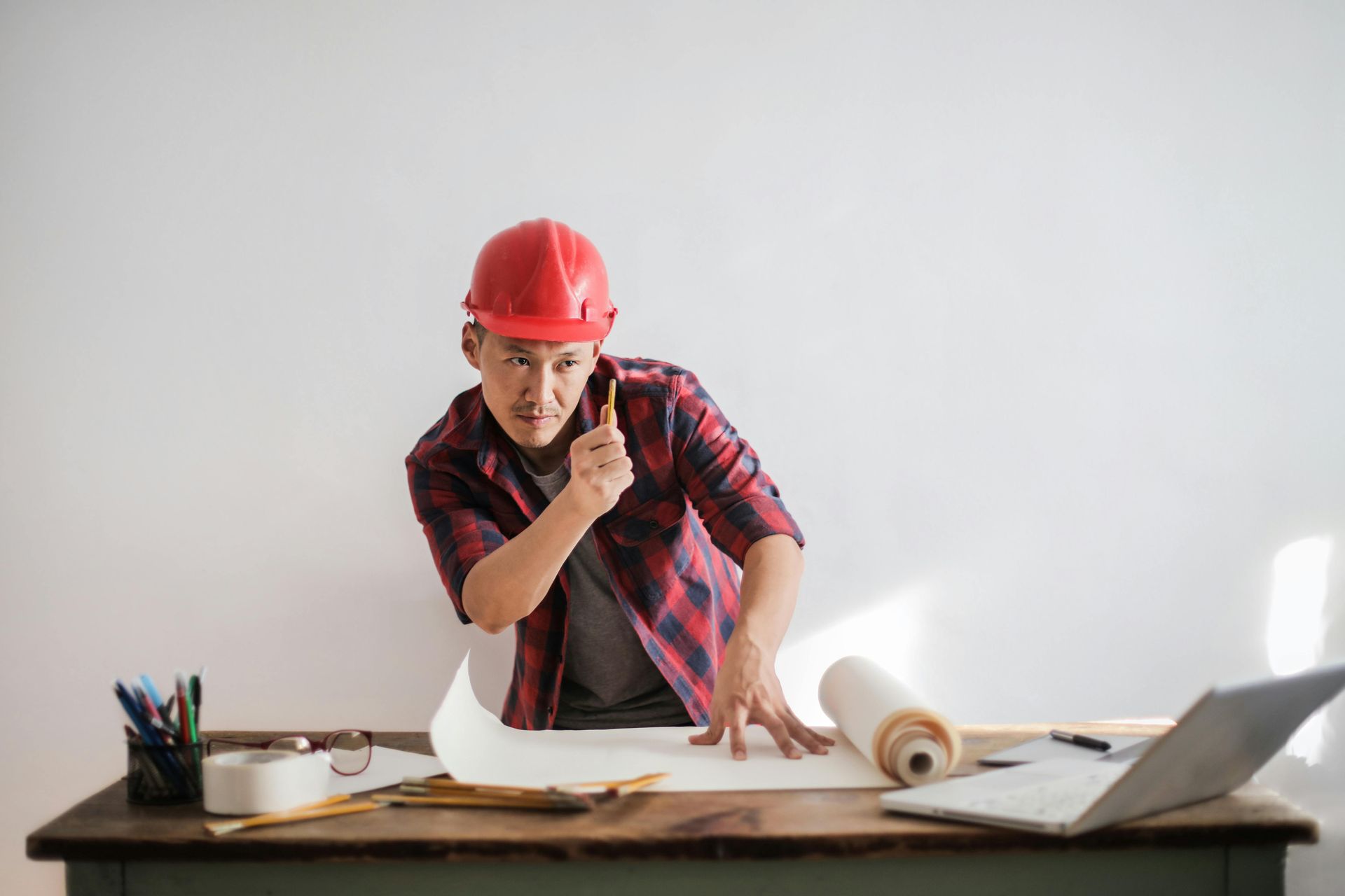 Man in red hard hat leans over blueprints, pointing pencil, at a desk.