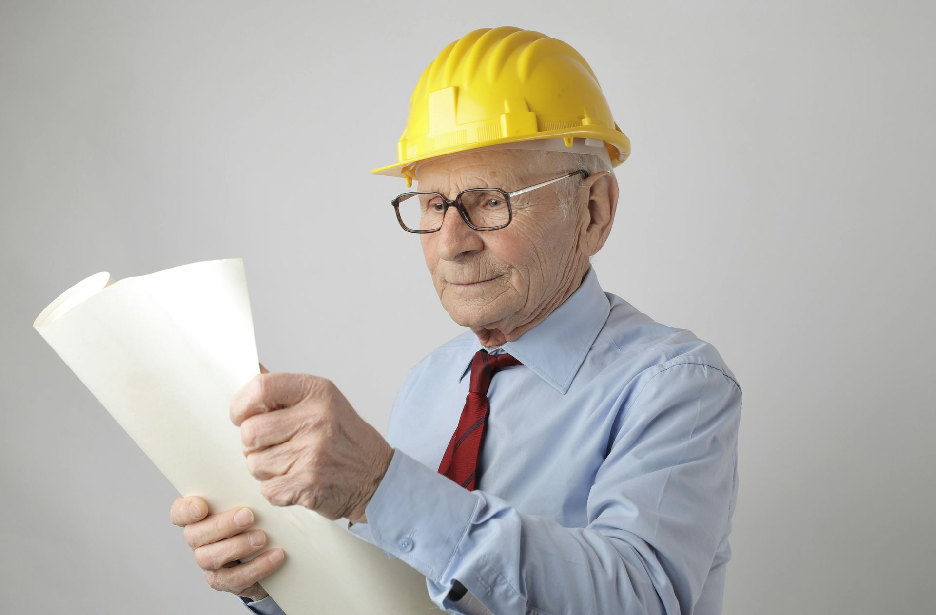 Older person in yellow hard hat and glasses, inspecting architectural plans.