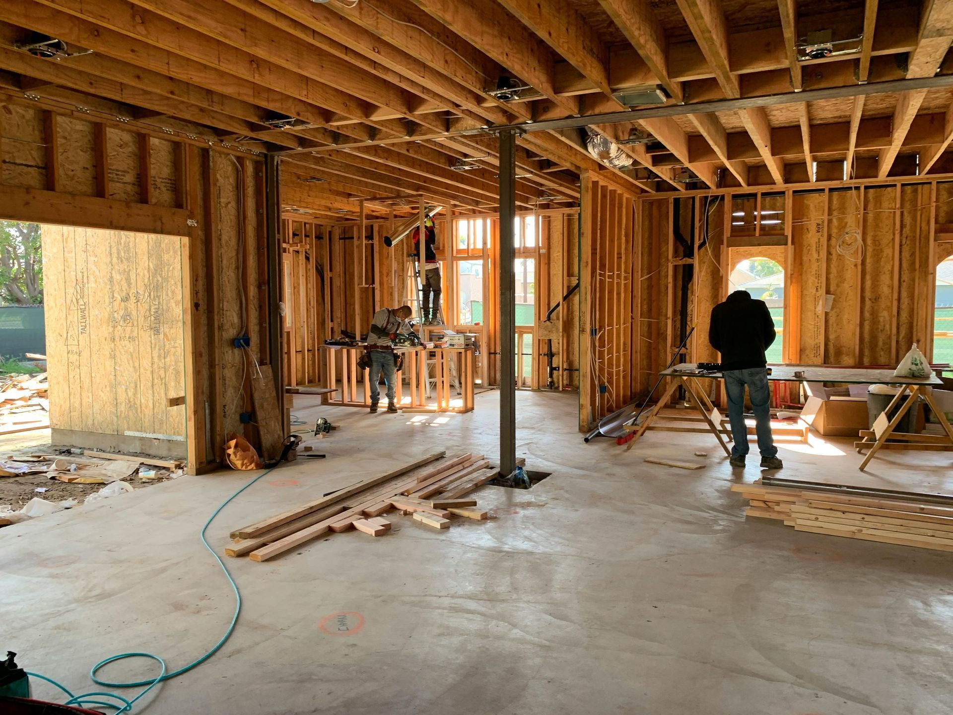 A construction site with wood framing, exposed ceiling joists, concrete floors, and two people working inside.