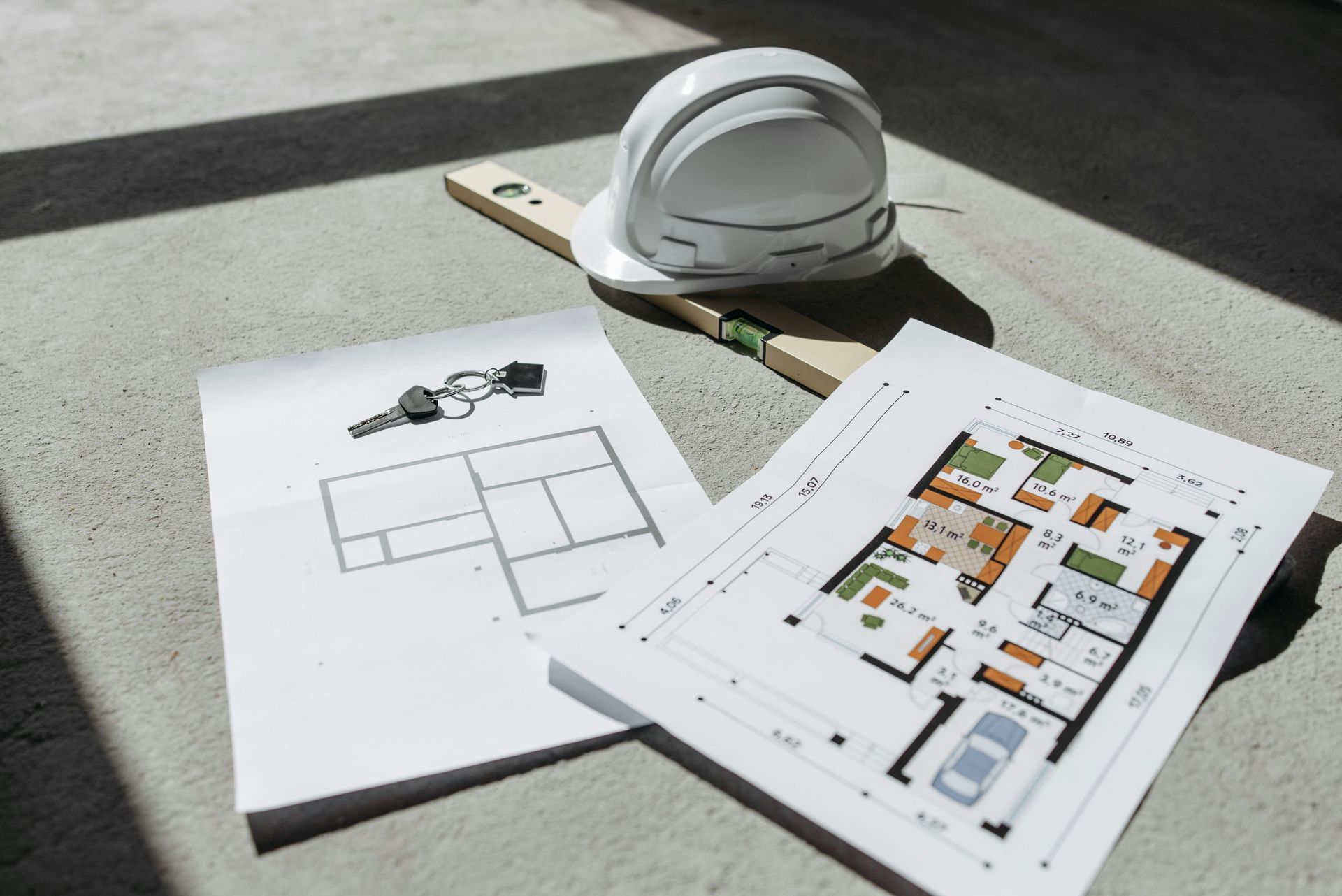 A white hard hat, a spirit level, a set of keys, and house floor plans resting on a concrete surface.